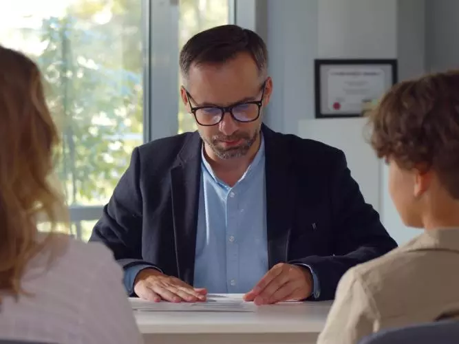 A male teacher sitting at a desk, talking to a mother and child who are sat opposite him with their backs to the camera.