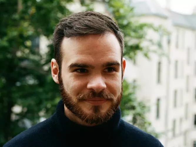 Man with dark brown hair and beard, wearing a black polo neck top