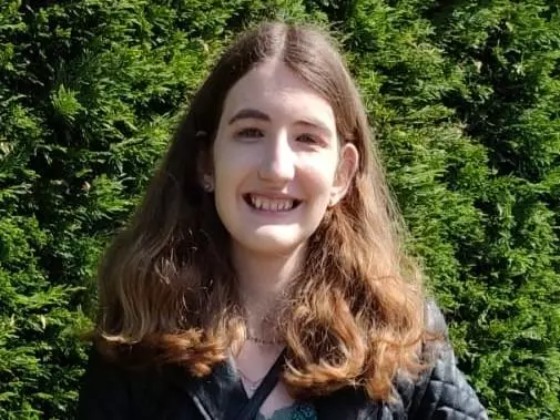 Chloe Brimicomb has shoulder-length brown hair and is wearing a blue jacket, standing in front of a green background