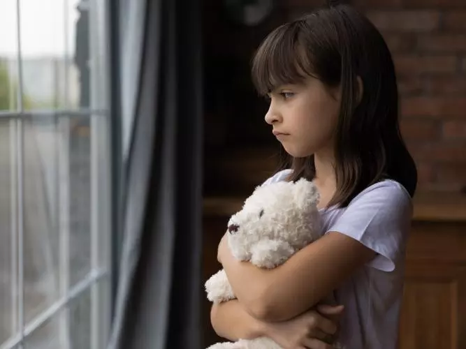 A young girl standing at a window, hugging a teddy bear. Credit: fizkes / Getty Images 1300371821