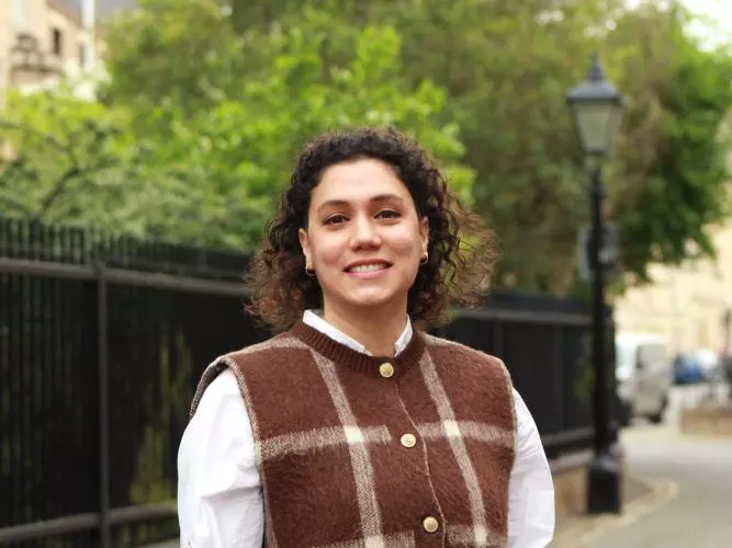 Elifcan has shoulder-length, curly dark hair. She is wearing a white shirt and a brown vest with a large white check. She is standing in front of a park fence, smiling. 