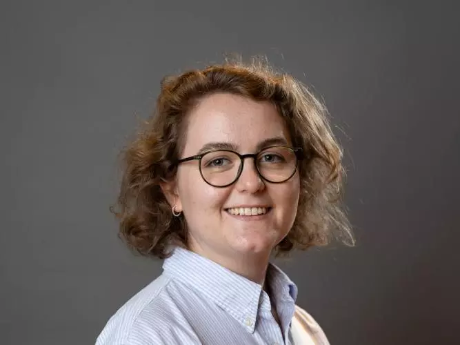 Leah Ennis waers a lab coat and glasses and is smiling in front of a grey background