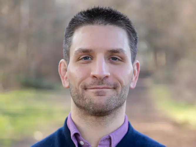 Lewis Anderson is wearing a dark blue sweater over a purple shirt, with short dark hair, looking directly at the camera.