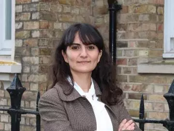 Merve is smiling and standing with her arms folded in front of a brown brick building. She has long dark hair and a fringe. She is wearing a white shirt and brown blazer. 