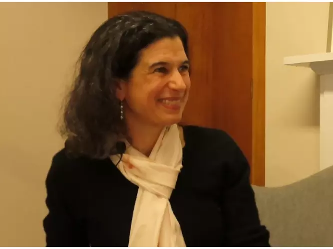 Professor Cluver has long curly black hair. She is wearing a black top and pale pink scarf.