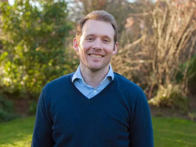 Alex Sutherland has short fair hair and is wearing a light blue shirt with a navy jumper. He is standing outside in a garden, smiling at the camera. 