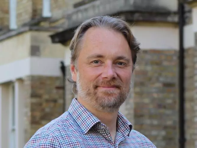 Mark Fransham is wearing a plaid shirt, with short blonde hair, a short bear and mostashe and smiling gentle at the camera.