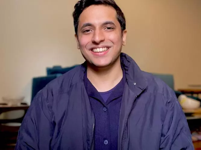 Ahsan smiles broadly at the camera. He has short brown hair and is wearing a navy blue knitted shirt and jacket. 