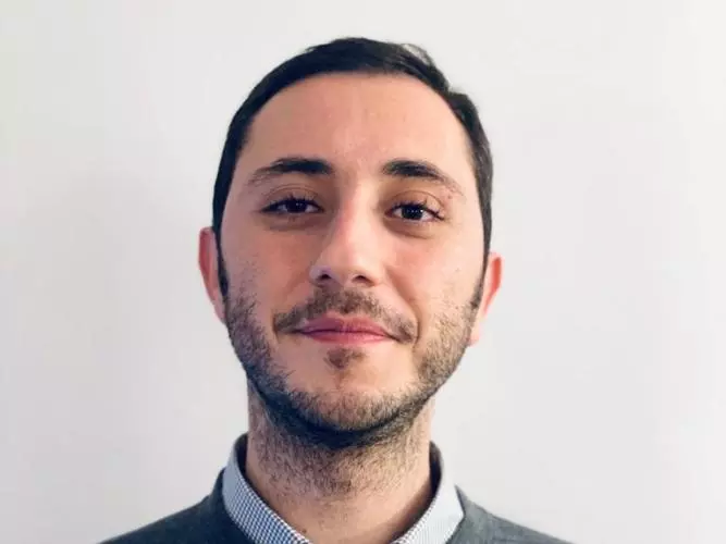 Dr Bedük has short dark hair. He is wearing a light blue shirt and grey jumper. 