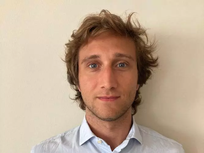 Enrico is standing in front of a wall looking at the camera. He has fair hair and is wearing a light shirt 