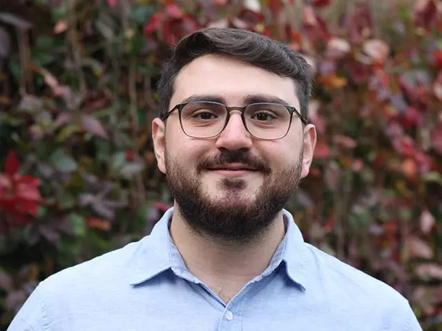 Ertugrul has short brown hair,  a brown beard and glasses. He's wearing a pale blue shirt