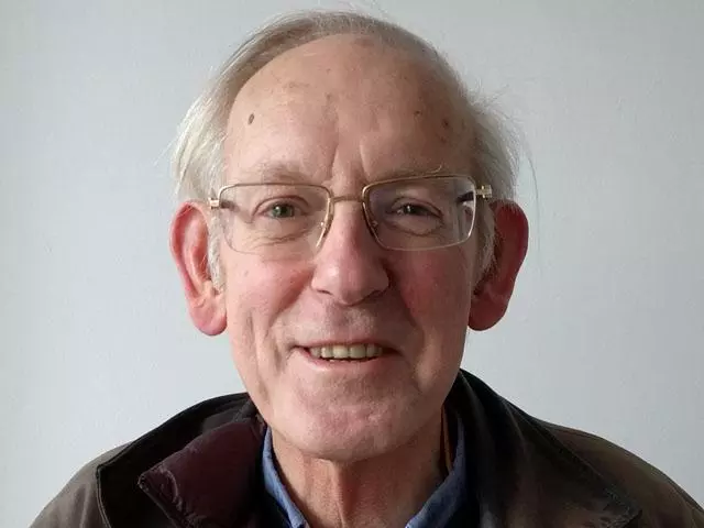Smiling man with short grey hair, bald head wearing a dark jacket and with gold rimmed glasses.