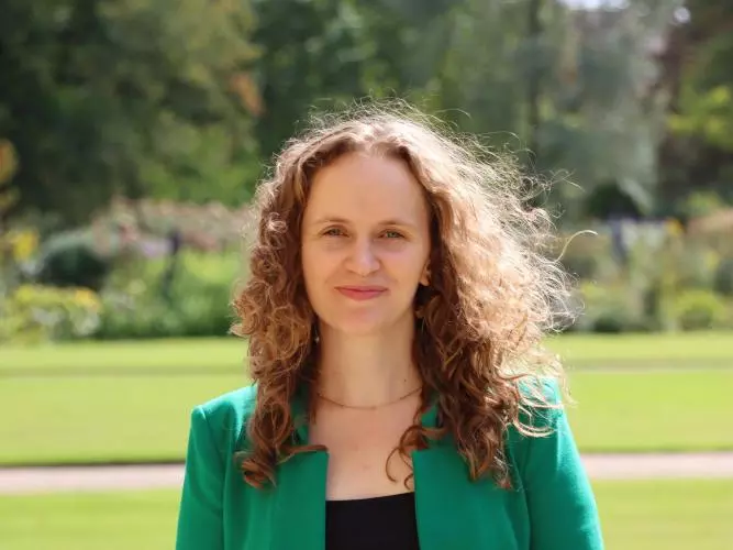 Jessica Lorimer has long curly brown hair. She is wearing a black dress and bright green blazer. She is standing outside in a large garden. 
