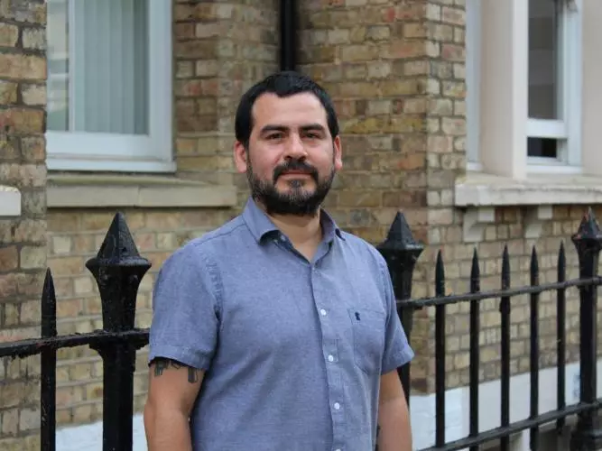 Dr Munoz has short black hair and black facial hair. He is wearing a short-sleeved blue shirt and standing outside a brick building. 