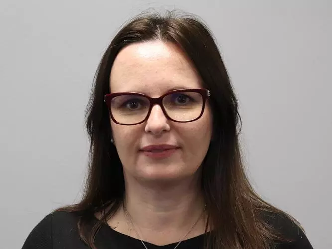 Woman with long brown hair, dark rimmed glasses and wearing a black top with necklace