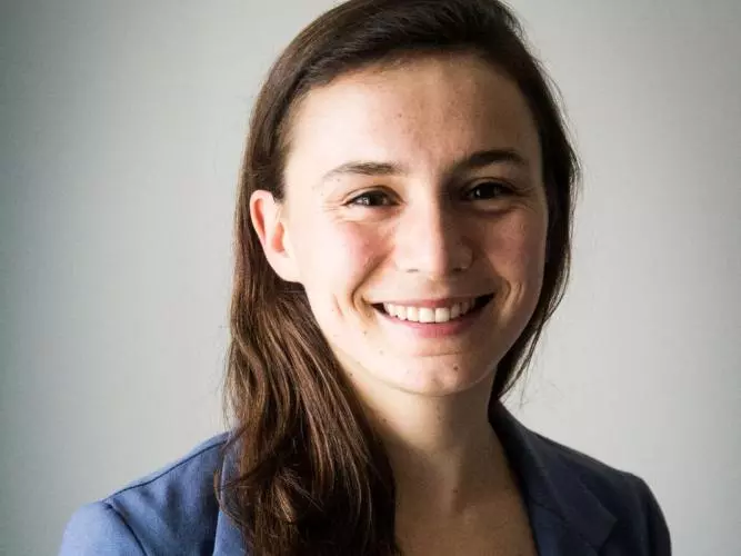 Madeline has long brown hair and brown eyes. She is wearing a blue jacket and white shirt. 