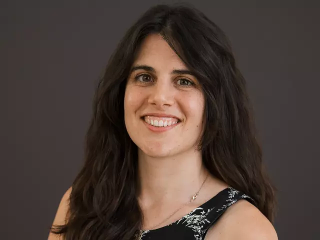 Woman with long brown hair, smiling at the camera and wearing a black dress with white pattern.