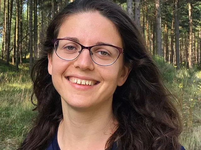 Yulia Shenderovich has long dark wavy hair, glasses and wearing a dark top. There is a background of trees behind her
