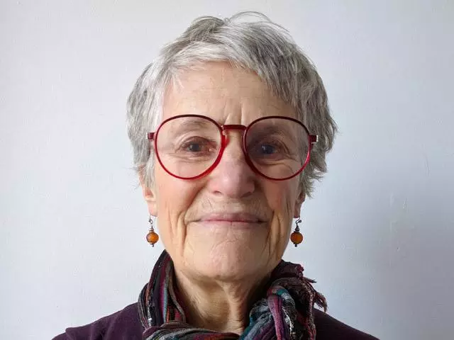 Woman with short grey hair, smiling and wearing red rimmed glasses and scarf around her neck.