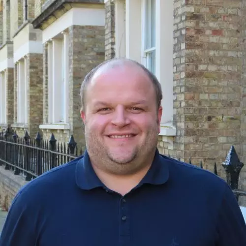 Man with broad smile wearing a dark blue shirt and jumper, and standing in front of brick buildings