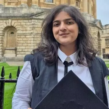 Shrutika Jha standing in front of the Bodleian Library. She has long wavy black hair and is in academic dress
