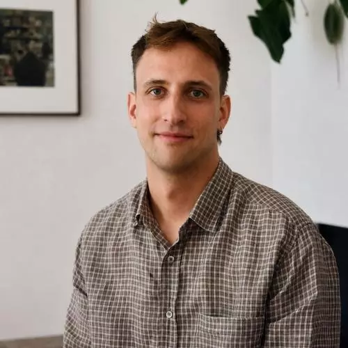Man with short brown hair wearing a brown checkered button-up shirt, seated indoors against a white wall with framed artwork and a leafy plant in the background