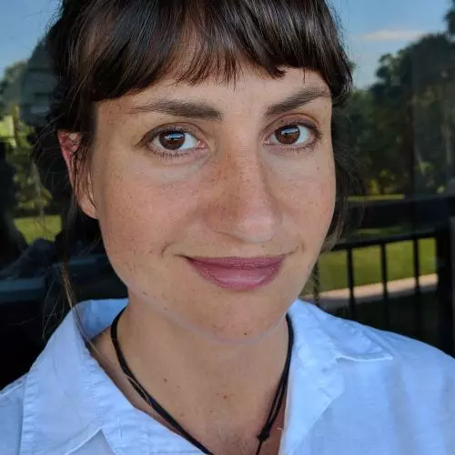 Smiling woman with short dark brown hair, wearing a light blue shirt and a black necklace. She is outdoors with trees and a railing in the background.