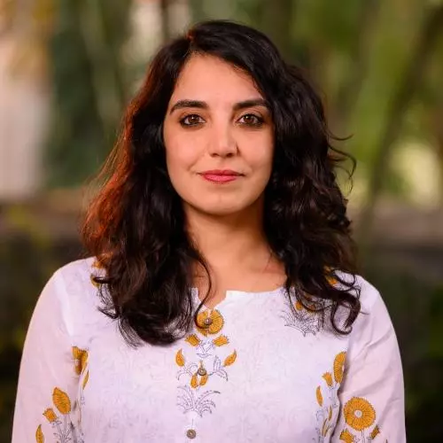Wearing a pink top with mustard flower patterns and shoulder-length brown wavy hair, Anita smiles gently at the camera.