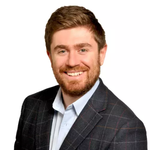Benjamin Scher has short ginger hair and a beard, wearing a blue shirt with a dark blue suit jacket, and smiling at the camera.