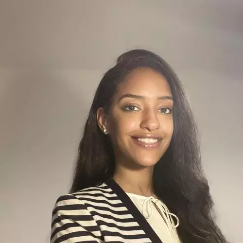 Bothaina has long black hair and is smiling at the camera. She's wearing a white top with a black and white horizontally-striped cardigan