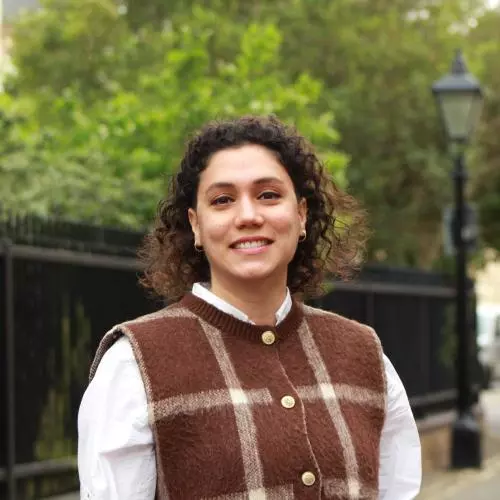 Elifcan has shoulder-length, curly dark hair. She is wearing a white shirt and a brown vest with a large white check. She is standing in front of a park fence, smiling. 
