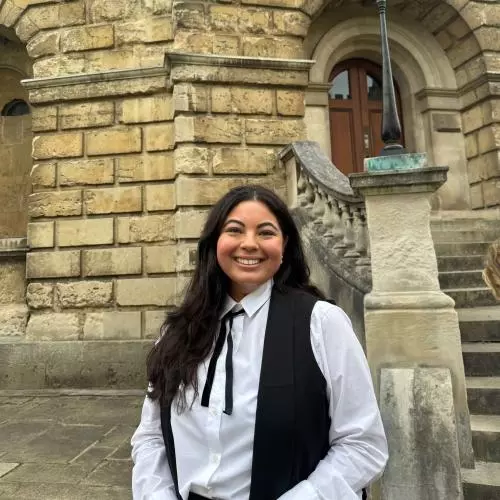 Lhori Webster is wearing academic dress and stands in front of a sandstone building in Oxford. 