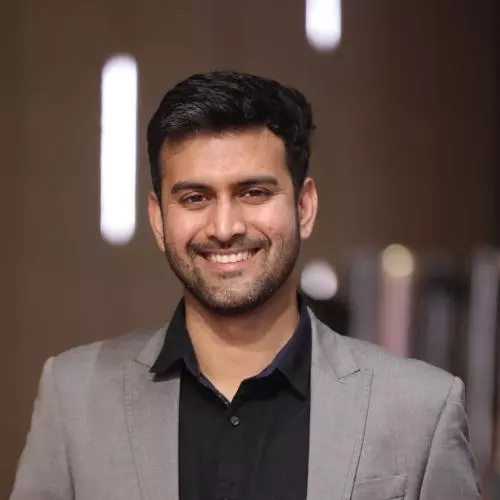 Sayyed has short black hair. He is wearing a black shirt and grey blazer