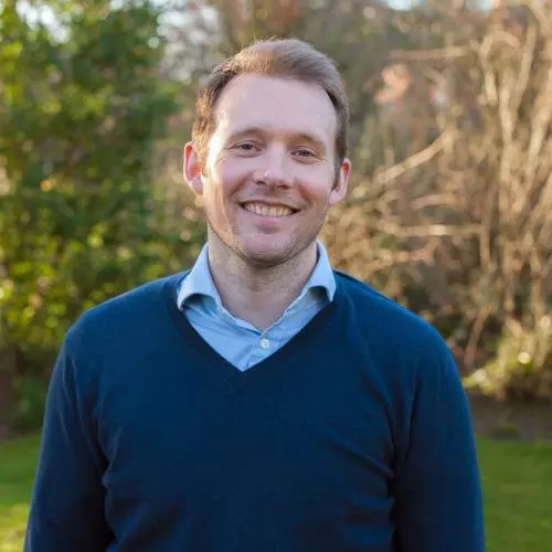 Alex Sutherland has short fair hair and is wearing a light blue shirt with a navy jumper. He is standing outside in a garden, smiling at the camera. 