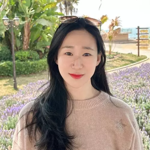 Lin Chen has long dark hair. She is standing in front of some flower beds and foliage and is wearing a beige knitted jumper. 