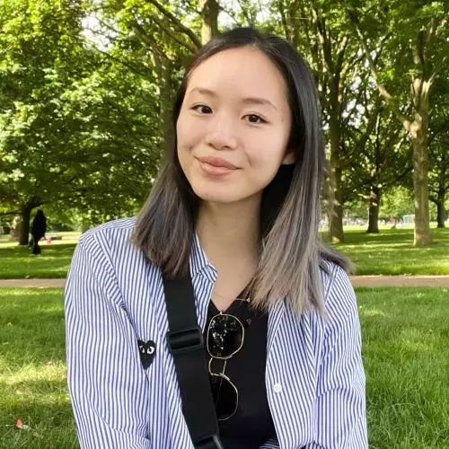 Victoria Tan has shoulder length dark hair, wearing a blue and white top and is sitting in a park.