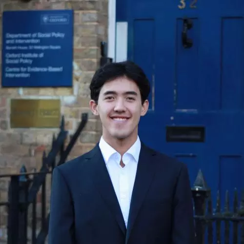 Xin Law-Gallagher stands, smiling, in front of the DSPI blue door. He wears a white shirt and dark blazer. He has short cropped dark hair. 