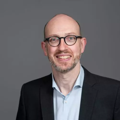 Arne Kuehl is wearing a suit and glasses smiling