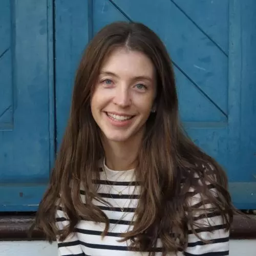 Bridget Steele has long brown hair and is wearing a black and white striped shirt. She is sitting on some outdoor steps in front of a blue door and smiling.