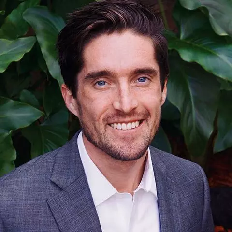 Man with dark hair who is smiling and wearing a white shirt and grey jacket. He is standing in front of a dark green hedge.