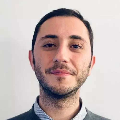 Dr Bedük has short dark hair. He is wearing a light blue shirt and grey jumper. 