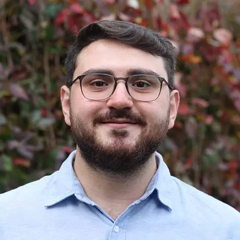 Ertugrul has short brown hair,  a brown beard and glasses. He's wearing a pale blue shirt