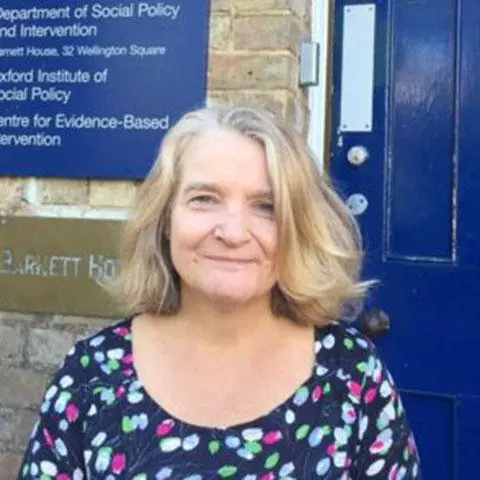 Smiling woman with light brown hair, wearing a patterned dress and standing in front of a dark blue door
