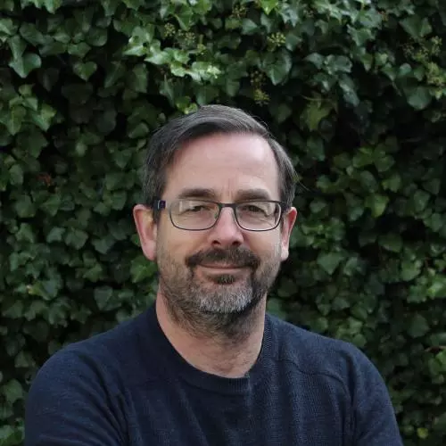 Gerry has short dark hair and a beard. He's wearing glasses and a navy blue jumper. He's standing in front of a hedge. 
