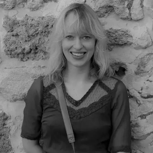 Janina has shoulder-length blonde hair and is smiling at the camera. She's wearing a dark-coloured top, and is standing against a stone wall
