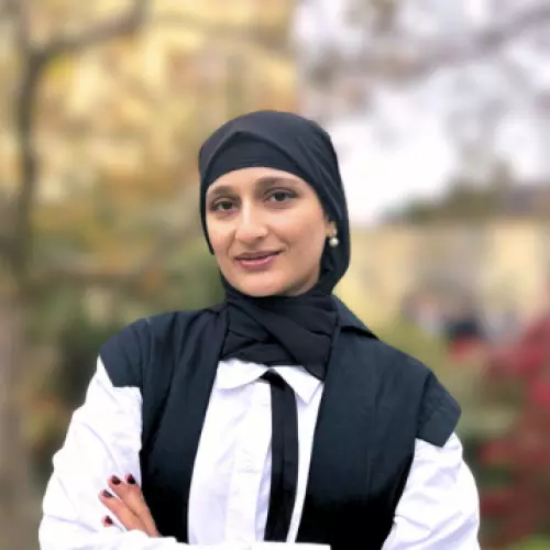 Kayanat Iltaf stands with folded arms wearing academic dress in front of an out-of-focus tree in the background. 