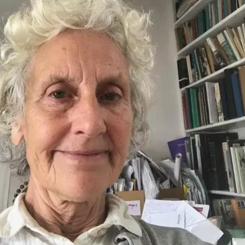 Dr Peretz has short curly white hair. She is sat in front of a large bookcase, smiling. 