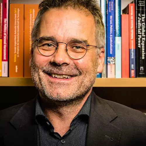 Martin Seeleib-Kaiser has short dark brown hair, glasses and is smiling at the camera. He's wearing a dark shirt and jacket and is sitting in front of a bookcase. 