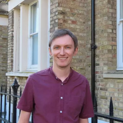 Robin has short fair hair and is wearing a short-sleeved burgundy shirt. He is standing outside in front of some light-brown brick buildings.