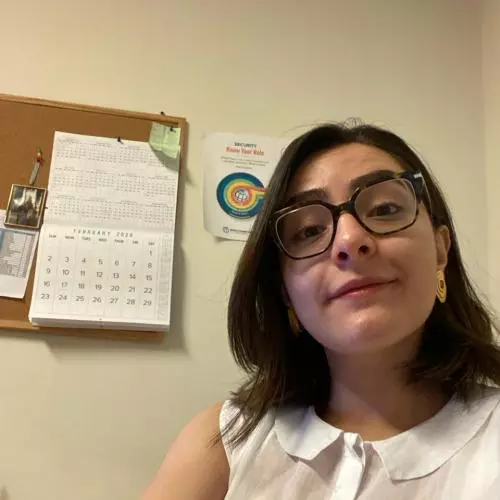 Nesma has shoulder-length dark hair, black-rimmed glasses and a white top. She is smiling at the camera